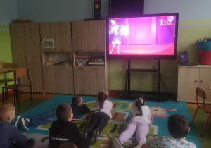 Dzieci leżąc na dywanie, oglądają na tablicy multimedialnej film edukacyjny o tradycjach andrzejkowych.