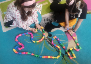 Uczennice siedząc na dywanie, budują najdłuższego węża z kolorowych klocków.