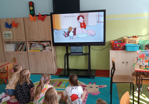 Uczniowie siedząc na dywanie oglądają na tablicy multimedialnej animowany film edukacyjny o pierwszej pomocy przedmedycznej.