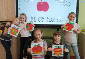 Dzieci na tle tablicy mulimedialnej prezentują wykonane przez siebie jabłka z papieru i kolorowej bibuły.