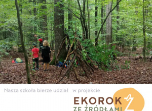Na zdjęciu widać dzieci spacerujące po lesie, mijające szałas z gałęzi.