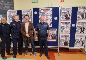Pan dyrektor z policjantem i dwiema policjantkami na tle plakatów promujących akcję Tydzień Przeciwdziałania Przemocy Rówieśniczej.
